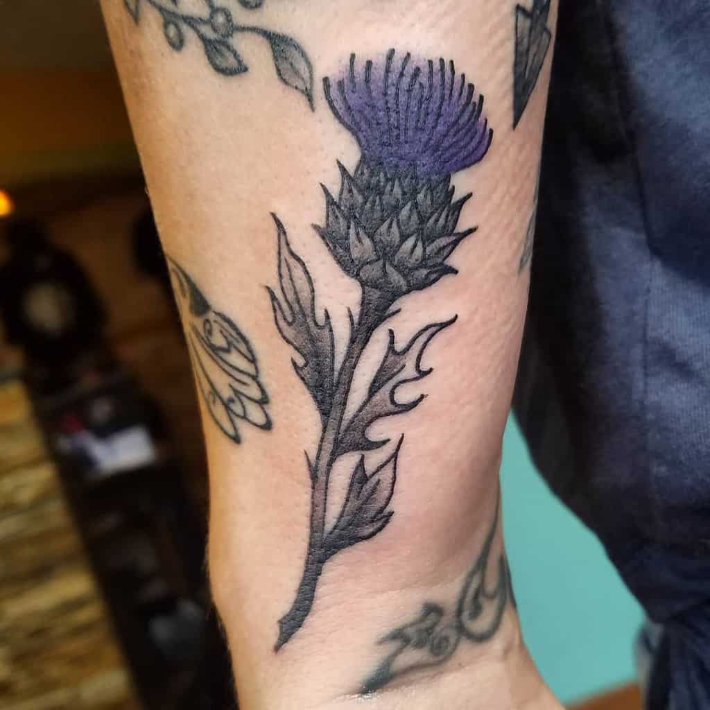 A stunning purple thistle tattoo blooms on a forearm adorned with other intricate designs