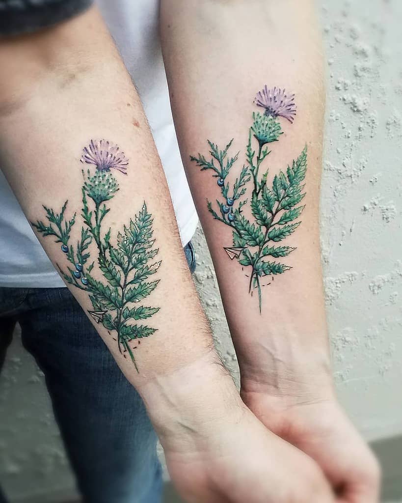 Two forearms with matching floral tattoos featuring green leaves and purple thistles