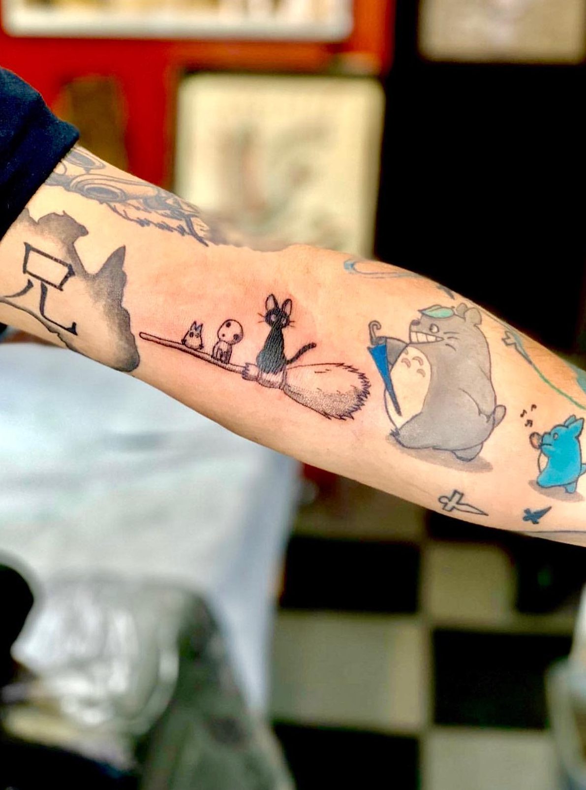 Playful Studio Ghibli tattoo on the forearm, featuring Totoro with an umbrella, Jiji on a broomstick with Kodama, and other small companions