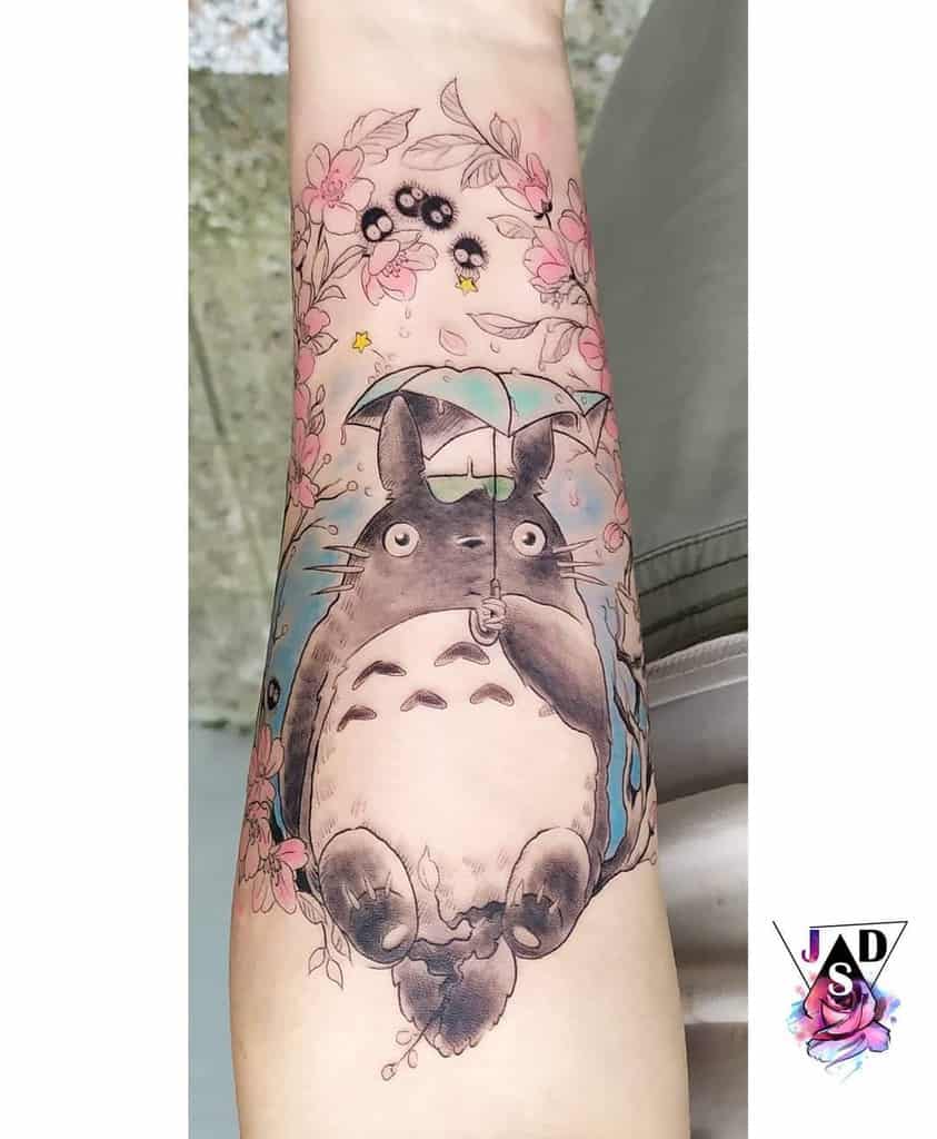 Beautiful Totoro tattoo on the forearm, featuring Totoro holding an umbrella, surrounded by pink cherry blossoms, soot sprites, and small yellow stars