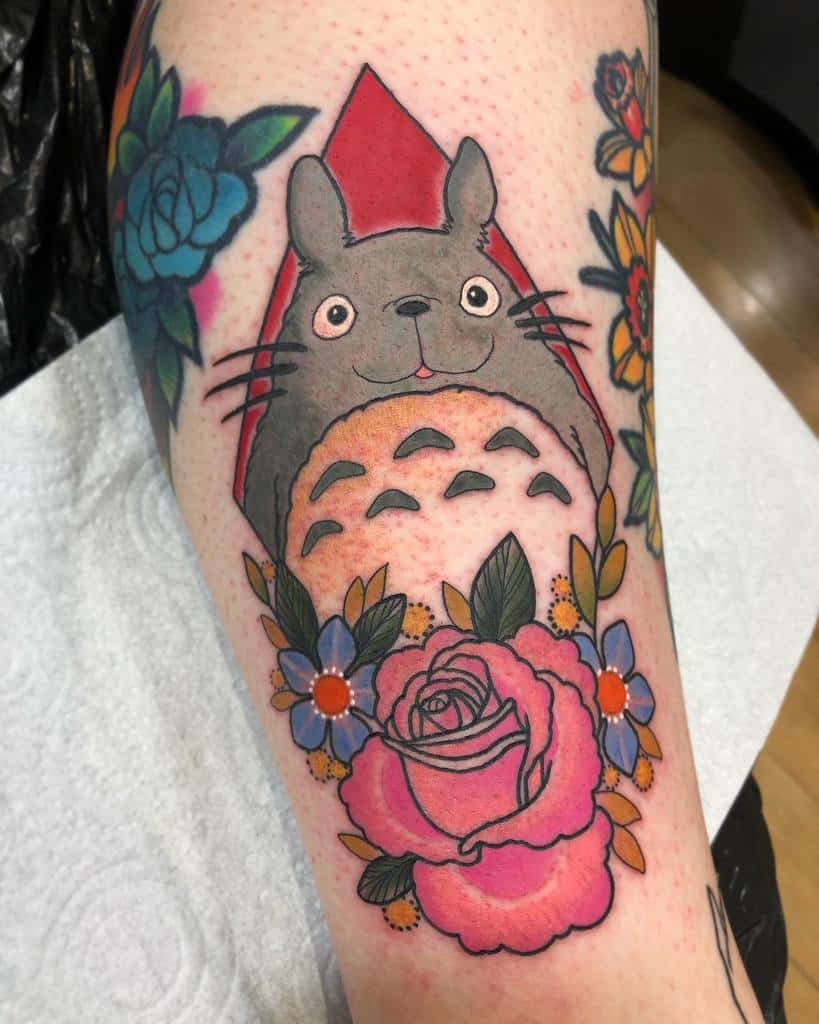 Bright Totoro tattoo on the arm, framed by a red triangle backdrop and surrounded by colorful roses, blue flowers, and leaves