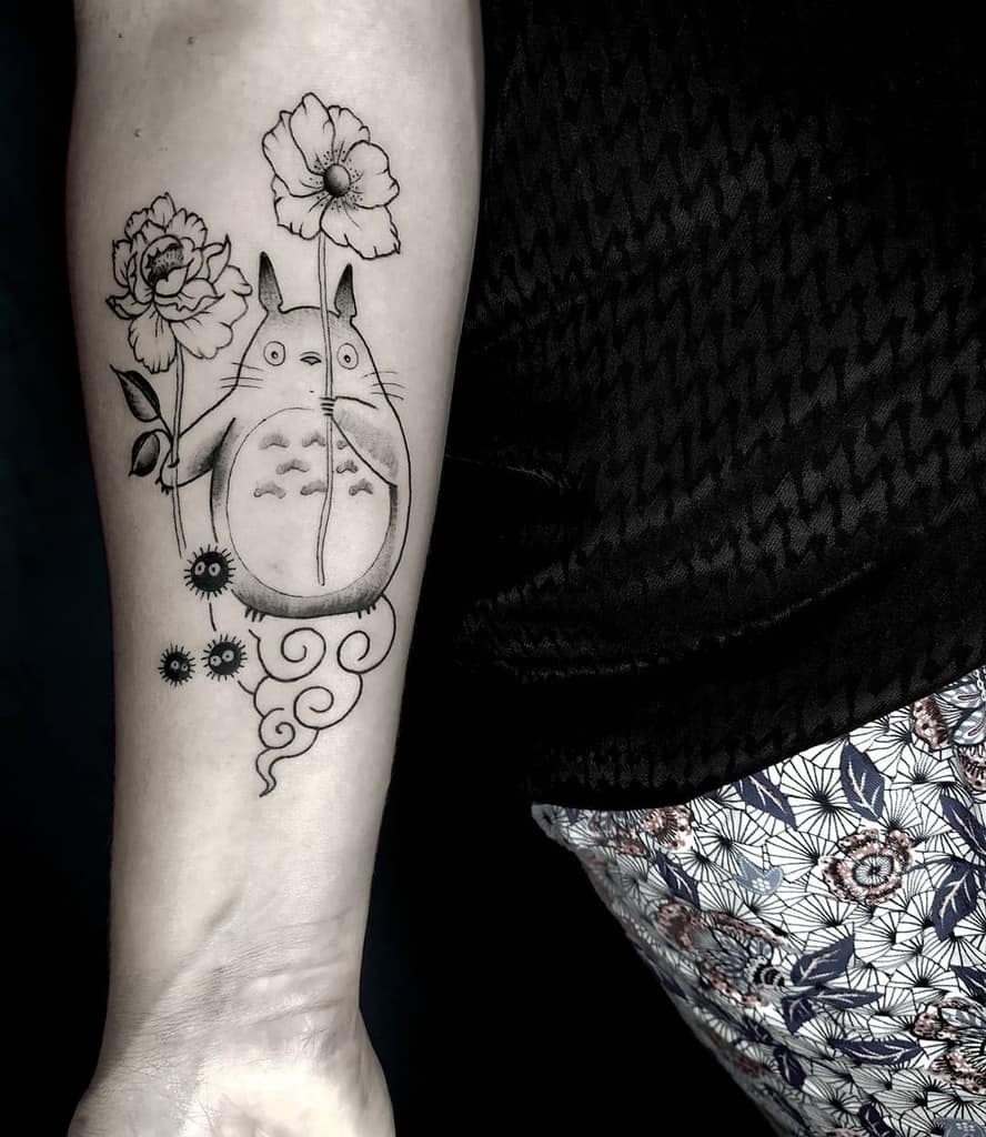 Elegant black and grey Totoro tattoo on the forearm, featuring Totoro holding tall flowers with swirling designs and soot sprites below
