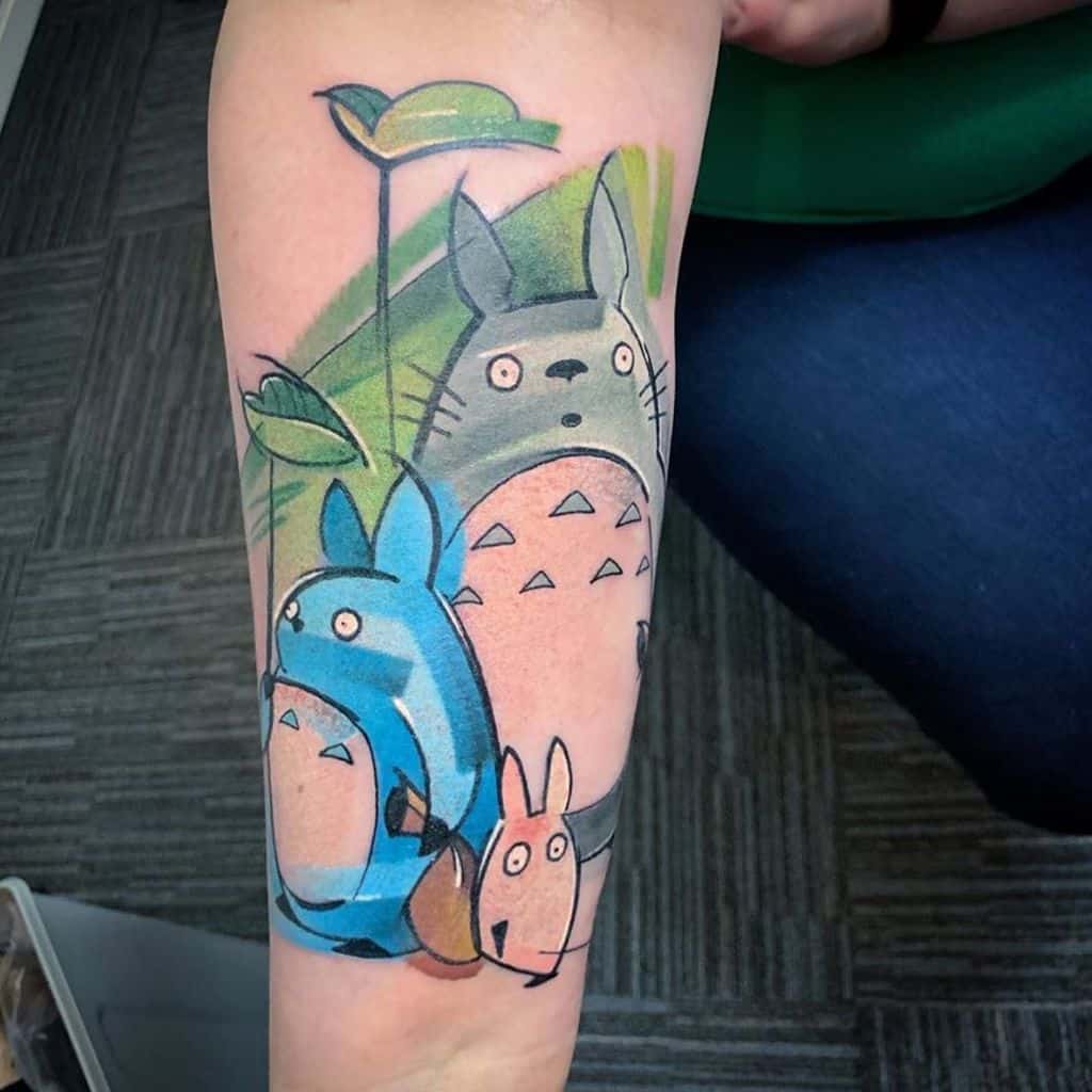 Colorful Totoro forearm tattoo featuring Totoro with his smaller companions, set against a green leaf backdrop