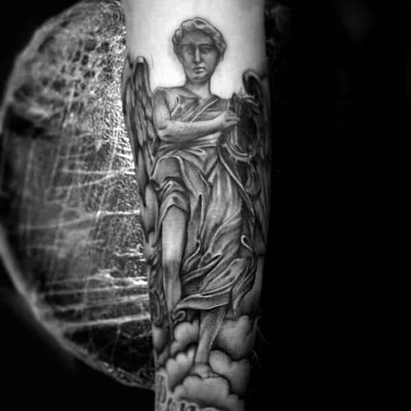 A black and grey tattoo on the forearm featuring an angel with flowing robes, surrounded by clouds, and detailed shading on the wings and figure.