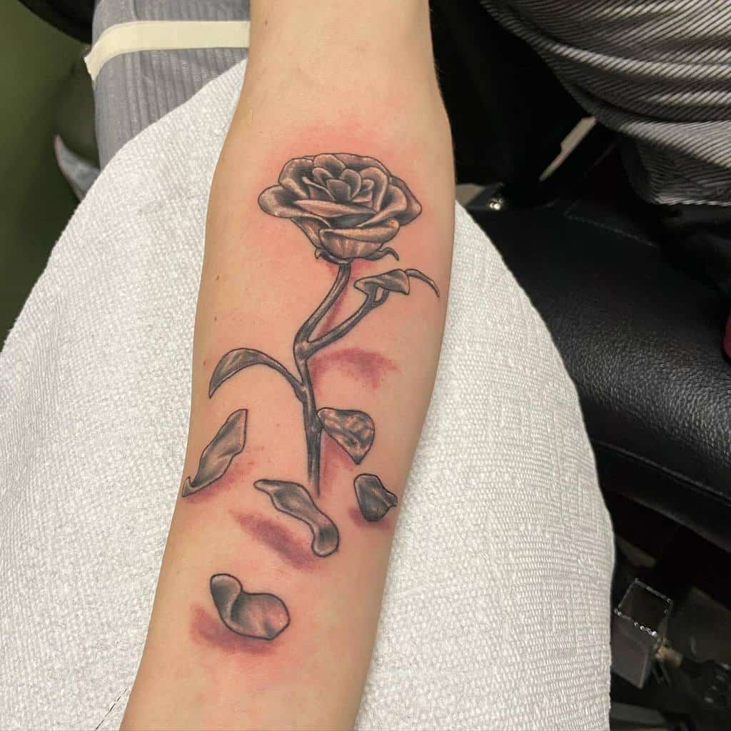 A detailed black and grey tattoo of a Beauty and the Beast rose, featuring falling petals and realistic shading on the stem and flower