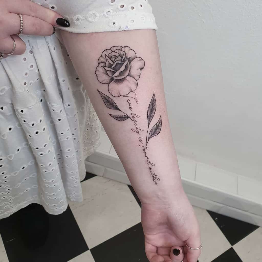 A black and white forearm tattoo of a rose intertwines gracefully with elegant text