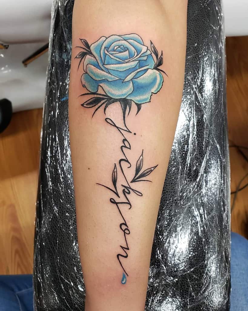 A vivid tattoo of a blue rose adorns the arm, elegantly entwined with the name "Jackson," while a delicate, tear-shaped droplet completes the poignant design
