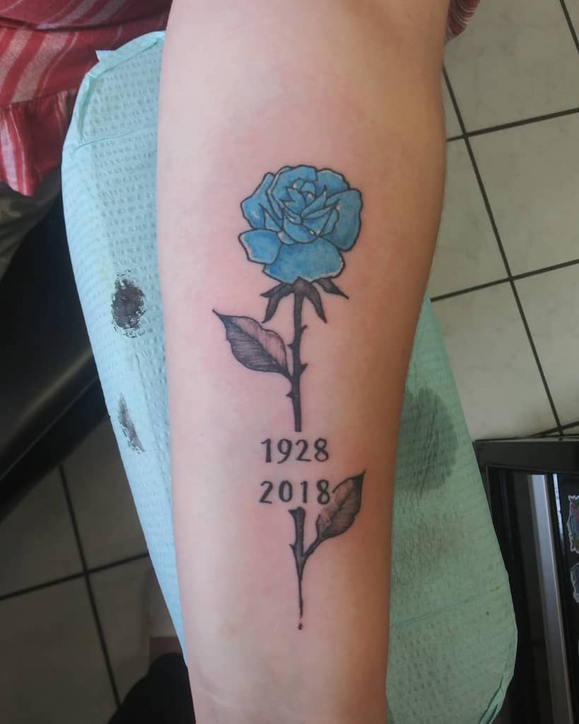 A striking tattoo of a blue rose adorns an arm, with the years "1928" and "2018" elegantly etched into its design. The vivid bloom stands out against a tiled background, capturing a poignant story within its petals