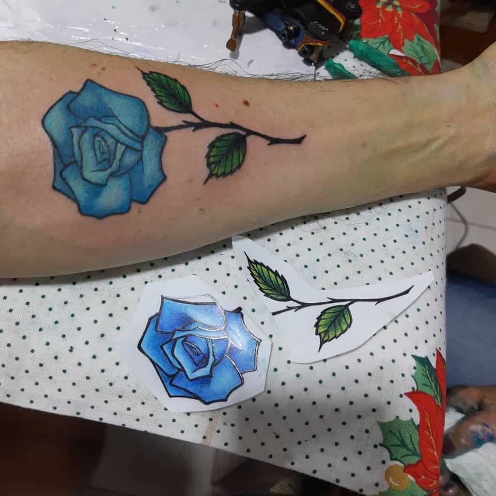 A colorful blue rose tattoo on the forearm, with detailed green leaves and a black stem, along with a design stencil placed beside it