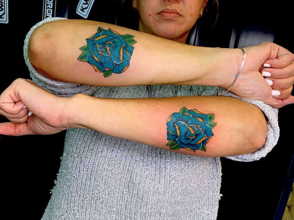 A matching pair of vibrant blue rose tattoos on both forearms, with bold green leaves and intricate detailing in orange and yellow accents