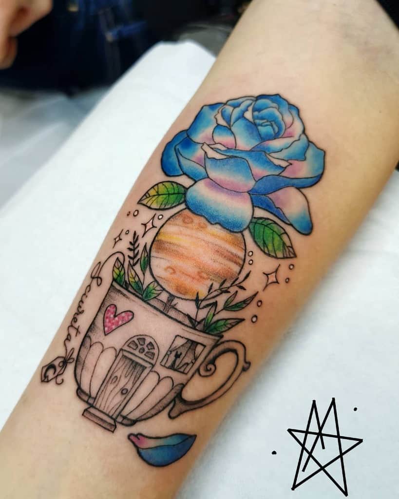 A whimsical tattoo on the forearm featuring a blue rose emerging from a teacup with a small planet, leaves, and stars, along with delicate details like a heart and bird