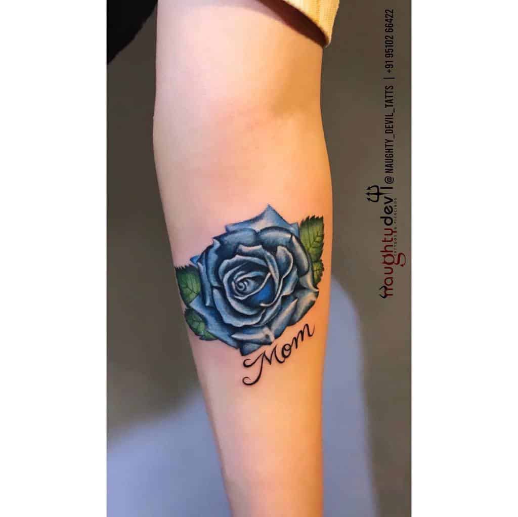 A stunning tattoo of a blue rose blooms gracefully on the arm, elegantly intertwined with the word "Mom"