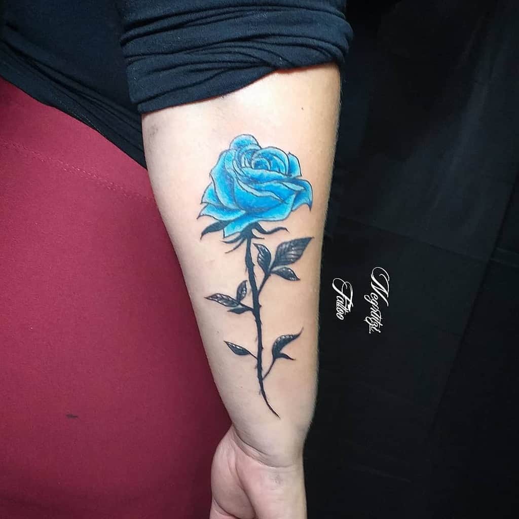 A stunning tattoo graces a person's forearm, featuring a blue rose with intricately detailed black leaves and stem