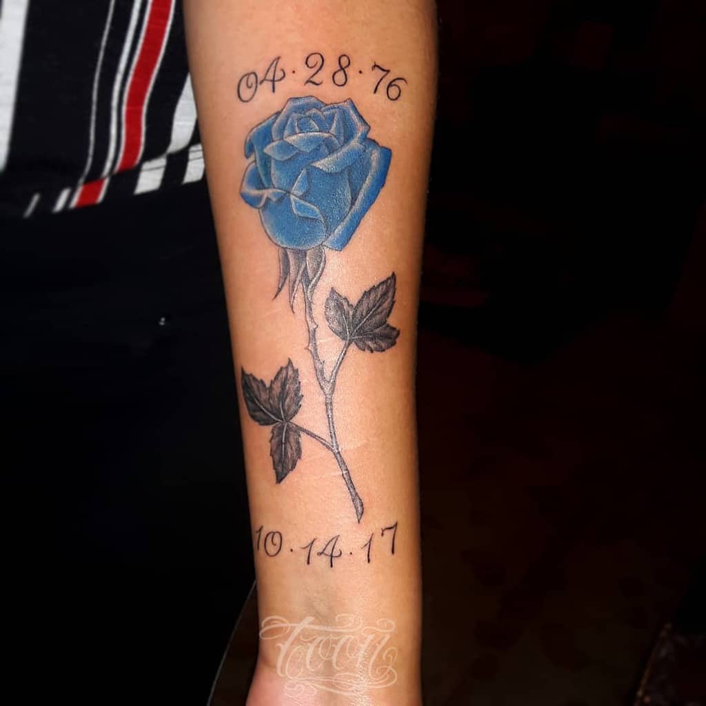 A tattoo featuring a stunning blue rose graces the forearm, accompanied by significant dates and a delicate handwritten script below
