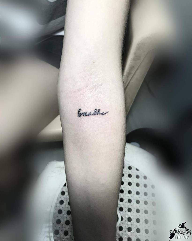 A close-up reveals an arm adorned with a graceful cursive tattoo of the word "breathe," serving as a gentle reminder to take in each moment