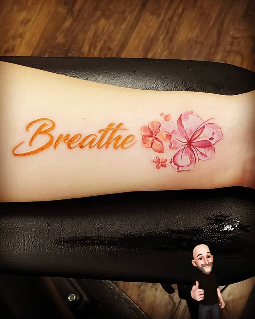 A graceful tattoo of the word "Breathe" adorned with delicate pink flowers elegantly graces an arm