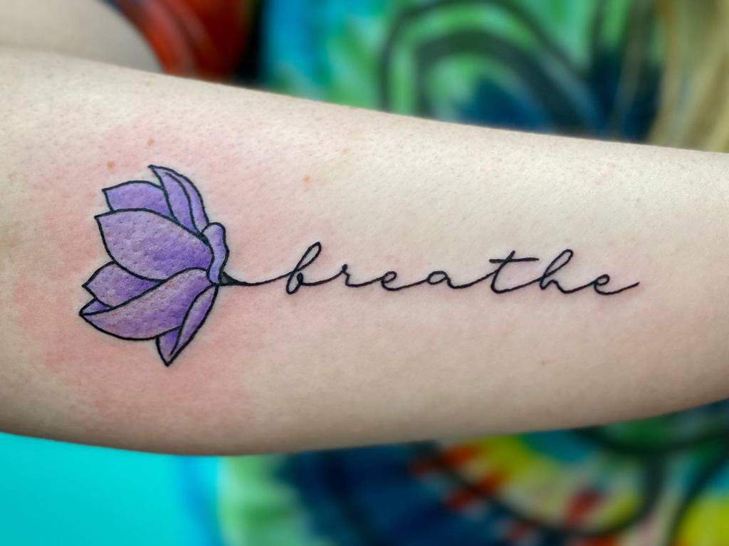 A beautiful tattoo of a purple flower graces the person's arm, elegantly intertwined with the word "breathe" in delicate cursive