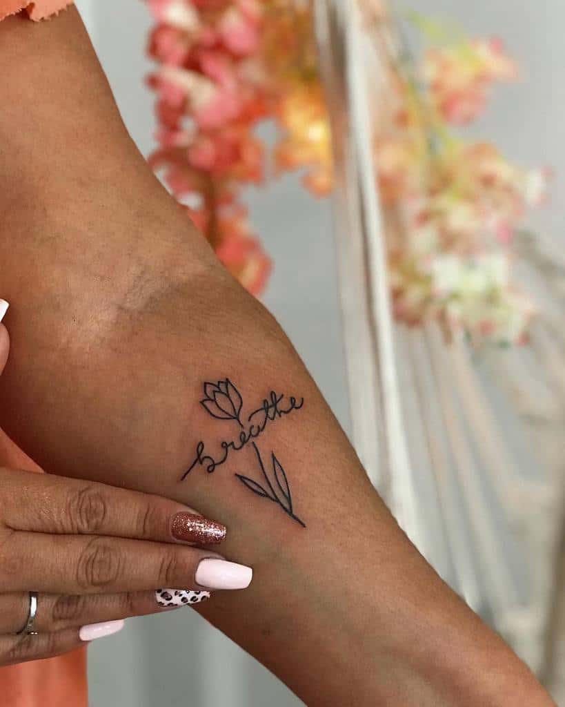 Arm adorned with a delicate tulip tattoo and the word "breathe" elegantly inscribed in cursive