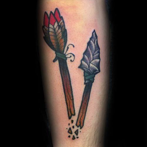 Traditional-style broken arrow tattoo with red-tipped feathers and a chipped stone arrowhead, surrounded by shattered fragments.