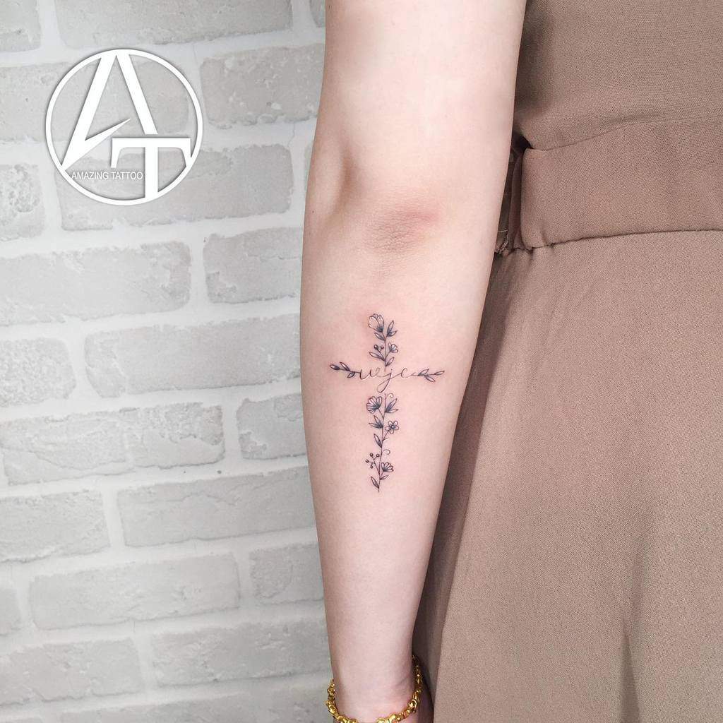 A delicate floral cross tattoo on the forearm, featuring intricate flowers along the vertical and horizontal lines with elegant cursive text across the middle
