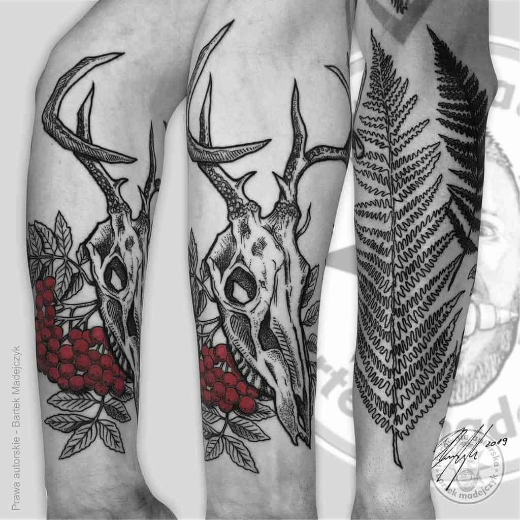 Black ink tattoos featuring a deer skull adorned with red berries, leaves, and a fern on an arm