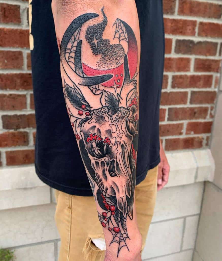 Intricate forearm tattoo featuring a deer skull with red accents, mushrooms, bat wings, and a crescent moon, blending dark and natural elements