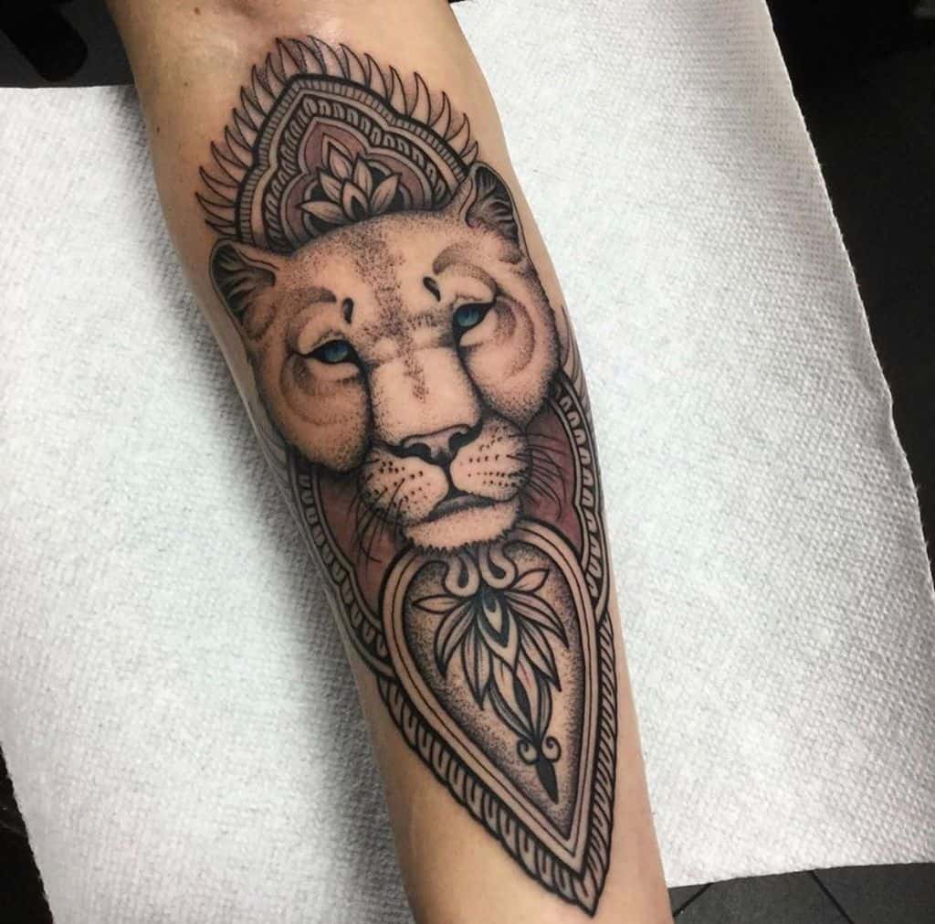 Intricate mandala-style lioness tattoo on the forearm, featuring bold black shading, geometric patterns, and striking blue eyes.