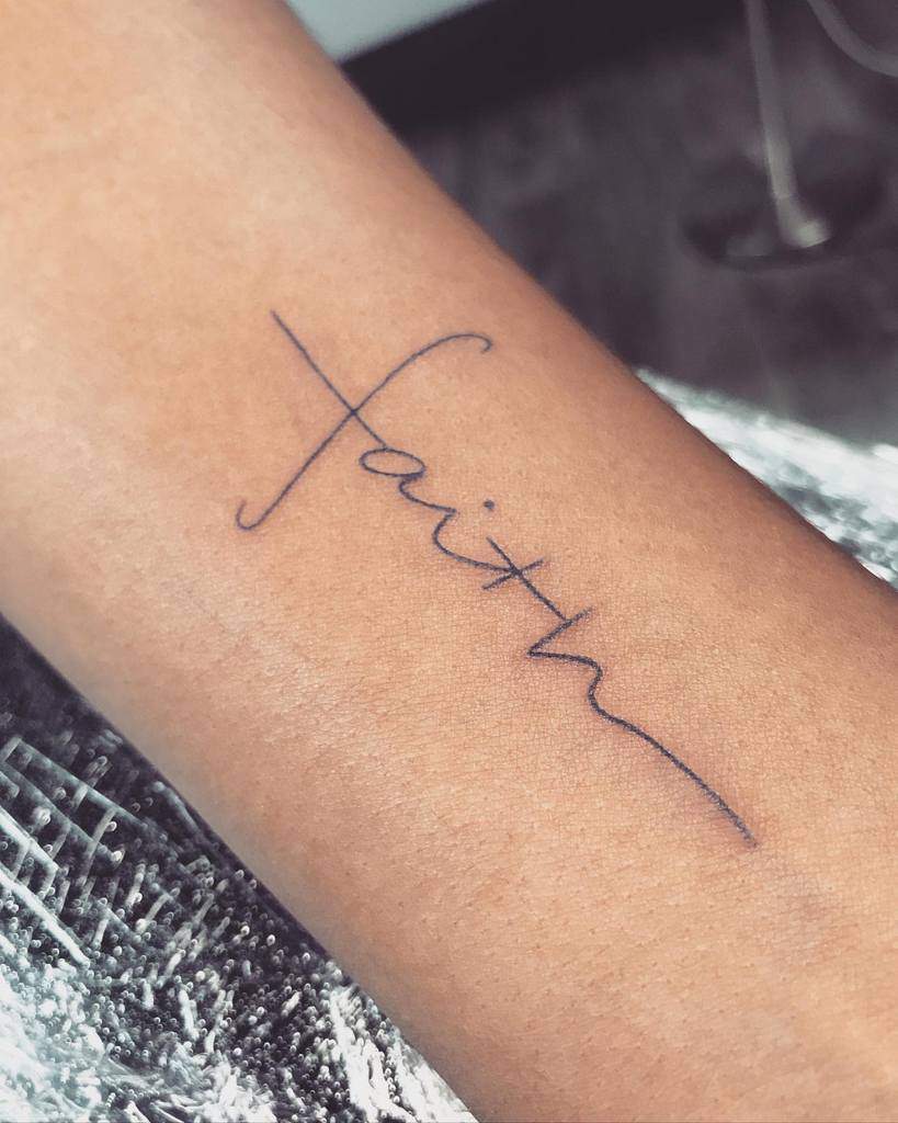 A delicate tattoo of the word "faith" elegantly drawn in cursive graces the arm, with a subtle cross intertwined, symbolizing unwavering belief and strength