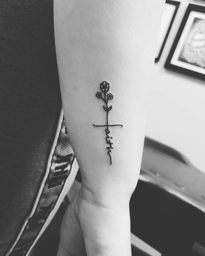 Black and white tattoo of a flower with the word "faith" in cursive intertwined with a subtle cross on an arm