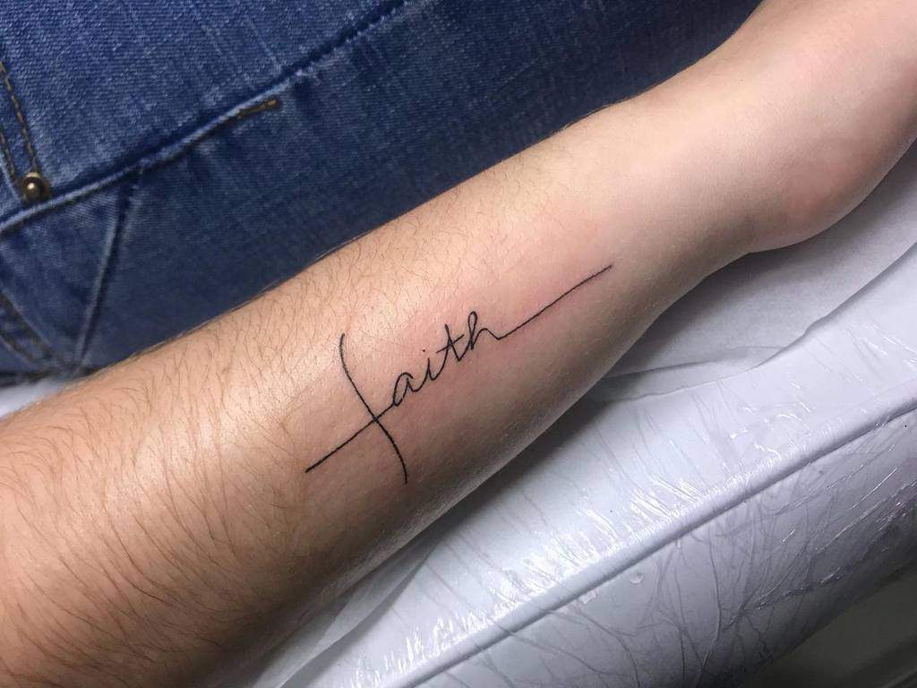 A tattoo of the word "faith" elegantly etched in cursive on a person's forearm, subtly adorned with a simple cross design