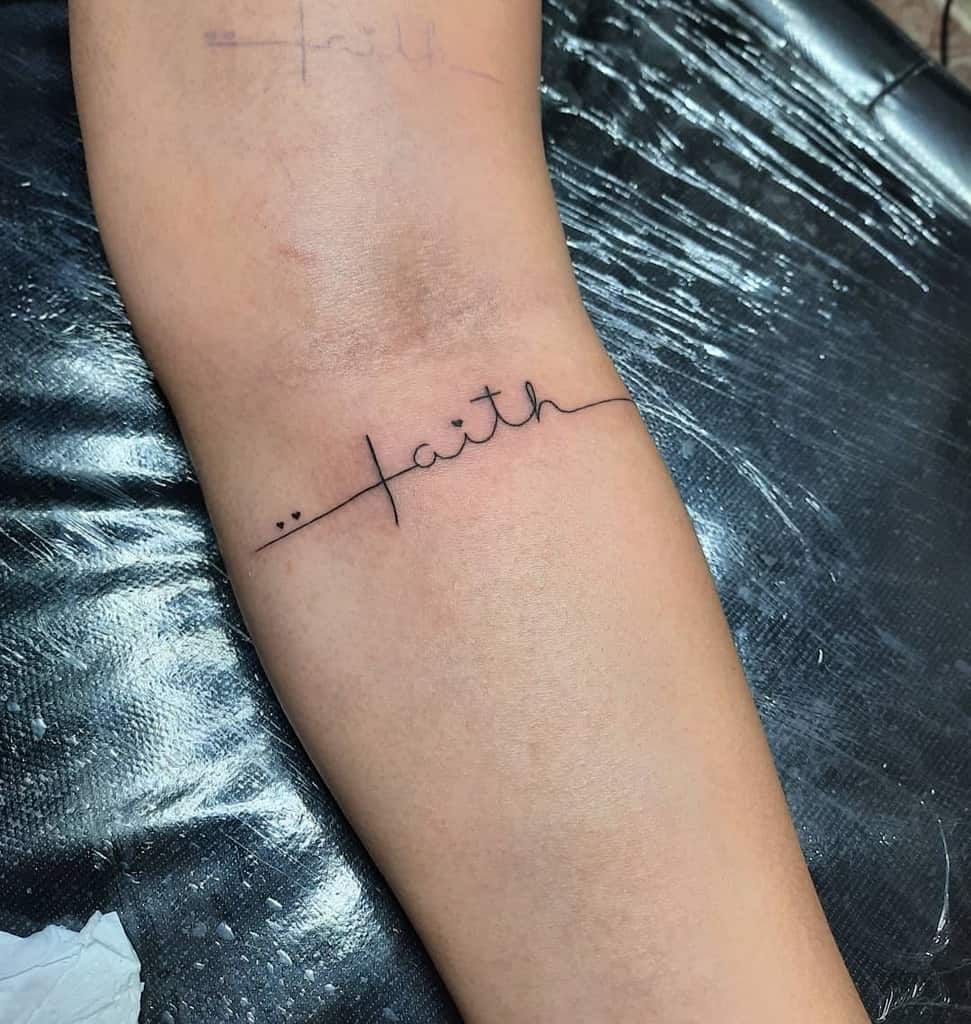 A graceful tattoo of the word "faith" in cursive elegantly adorns the arm, with a delicate cross integrated into the design, adding an extra layer of meaning and spirituality