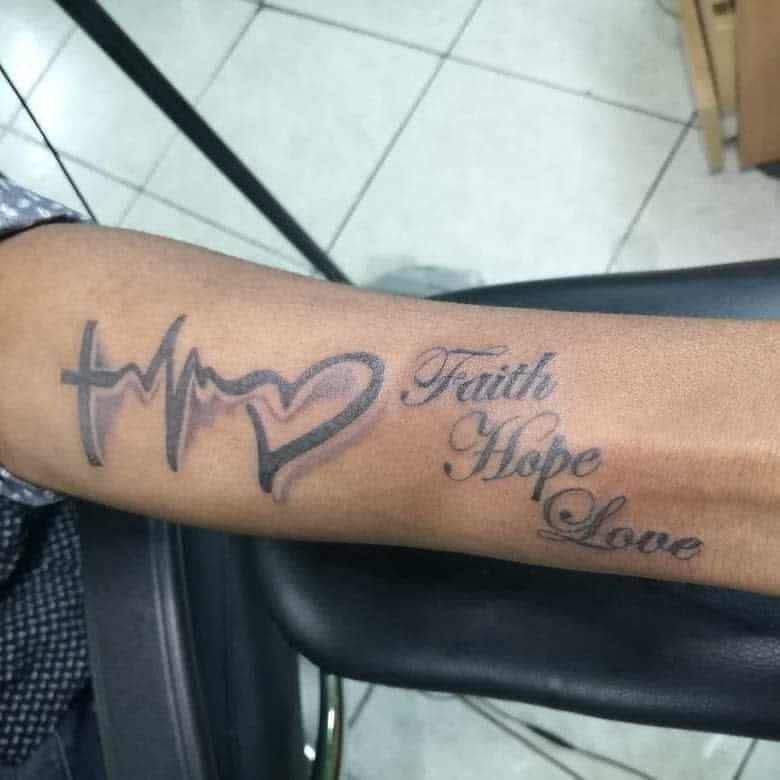 Forearm tattoo with a cross, heartbeat, heart, and the words "Faith Hope Love" seamlessly woven in cursive