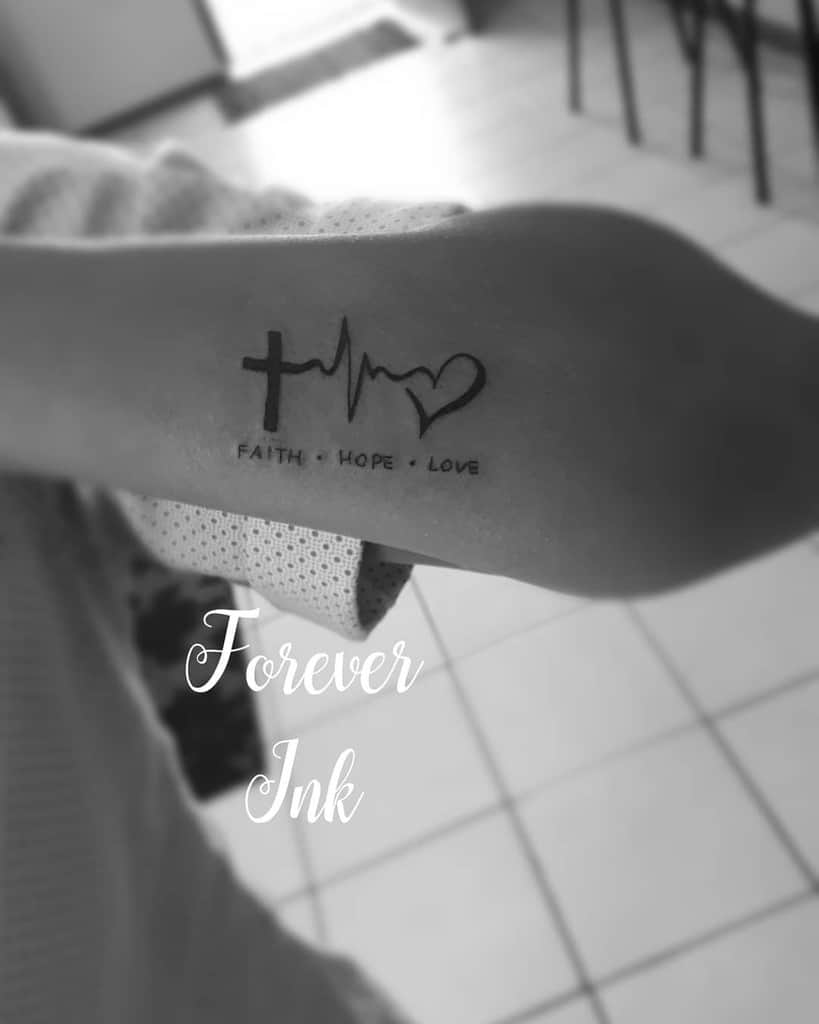 A forearm tattoo beautifully intertwines a cross, heartbeat, and heart design. The black ink inscription reads "Faith + Hope + Love," symbolizing a harmonious blend of devotion, optimism, and affection