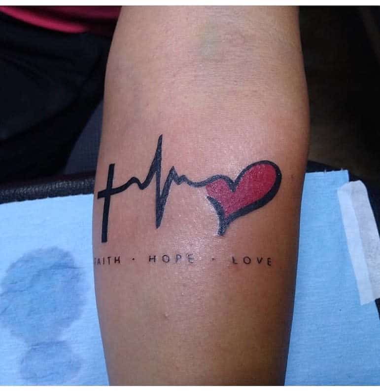 Tattoo of love, hope, and faith intertwined with a cross, heartbeat line merging elegantly into a vibrant red heart on an arm