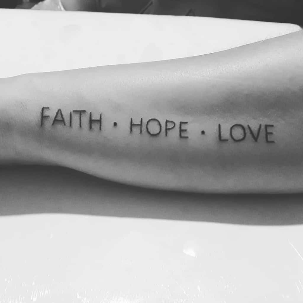 A grayscale image showcases a tattoo on an arm, elegantly inscribed with the words "FAITH HOPE LOVE" in bold black ink, capturing the essence of unwavering belief and affection