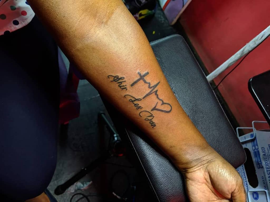 A forearm tattoo featuring a cross, heartbeat line, and heart symbol followed by cursive text 