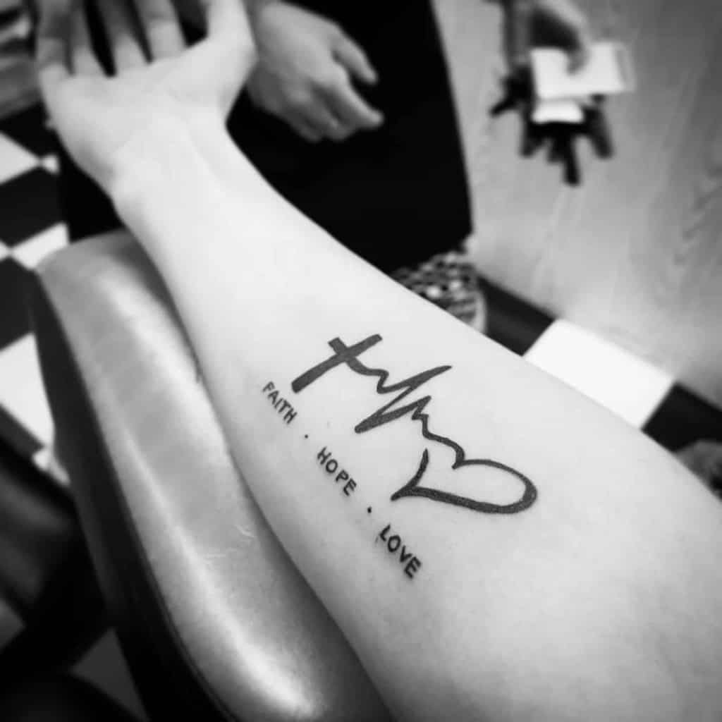 Forearm tattoo featuring a black ink cross, heartbeat line, and heart, beautifully complemented by the words "Faith," "Hope," and "Love"