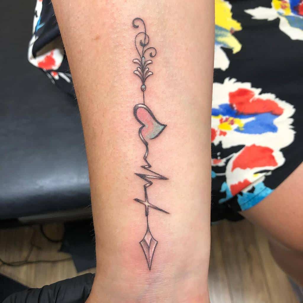 A forearm tattoo featuring a heart symbol, a decorative flourish at the top, and a heartbeat line with a cross integrated, leading to a geometric arrow design at the bottom