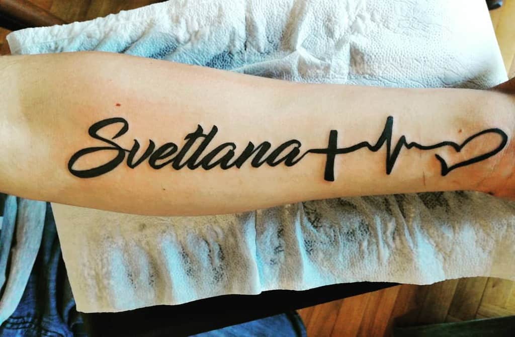 Forearm tattoo with the name 'Svetlana' in cursive script, followed by a cross, heartbeat line, and heart symbol