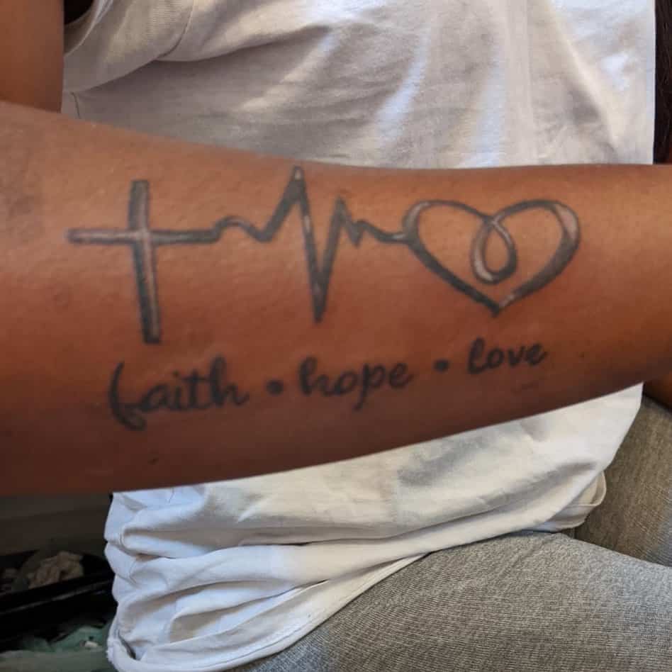 Tattoo on an arm with a cross, heartbeat, and heart symbols, encapsulating the words "faith • hope • love" in elegant script
