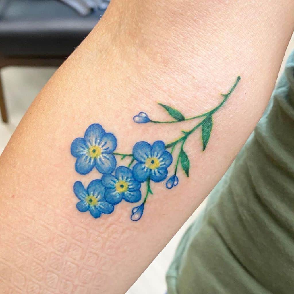 Tattoo of delicate blue forget-me-nots on an arm, complete with a graceful green stem and vibrant leaves