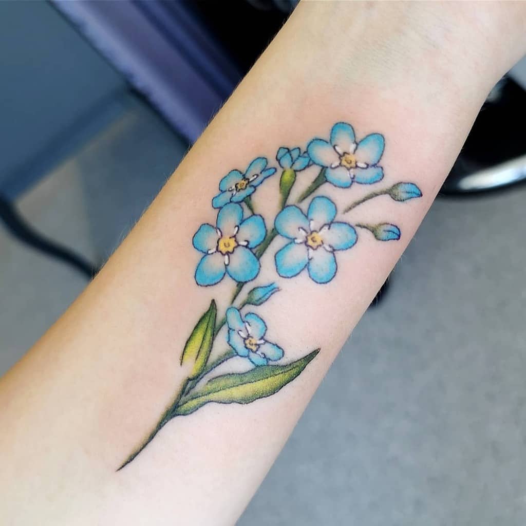 A tattoo of blue forget-me-not flowers with lush green leaves graces a forearm