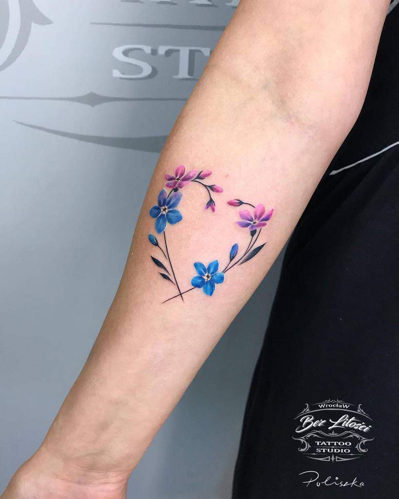 This tattoo features a delicate design of forget-me-not flowers in shades of pink and blue, forming a heart shape on the forearm, with soft watercolor shading and intricate line work