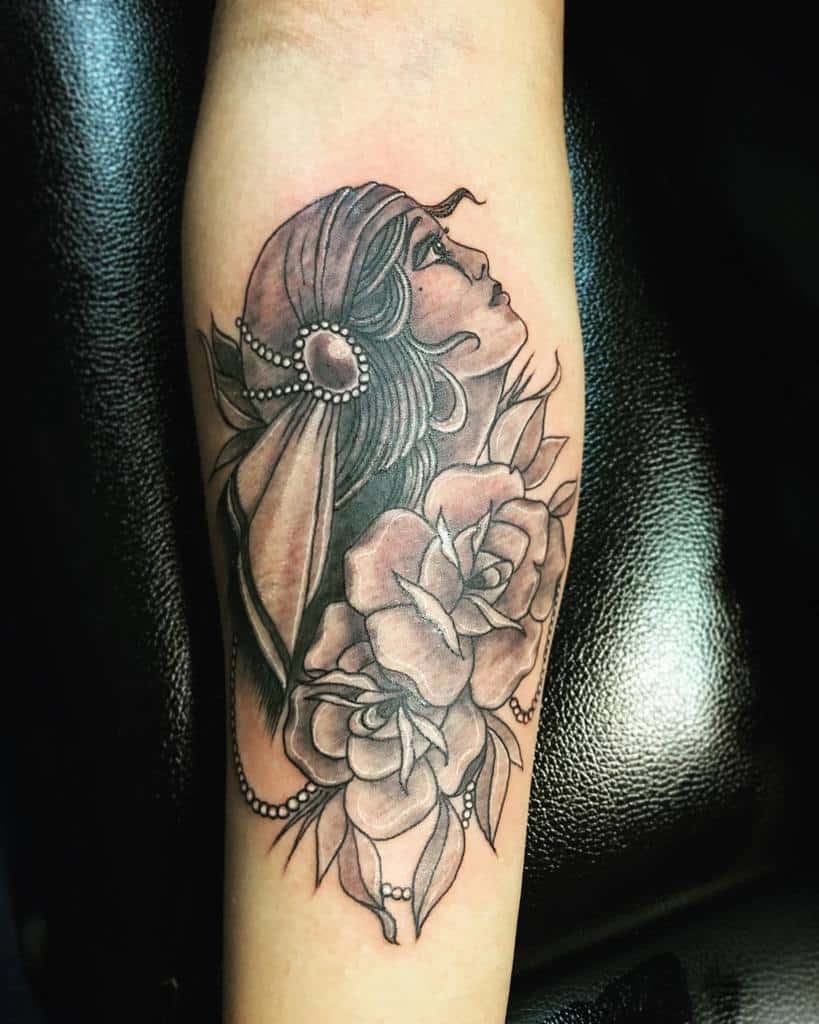 A black and gray tattoo of a gypsy woman with a headscarf and pearls, surrounded by roses, on the forearm