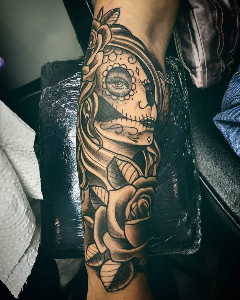 A gypsy-inspired tattoo of a Day of the Dead style face adorned with vibrant roses graces the forearm