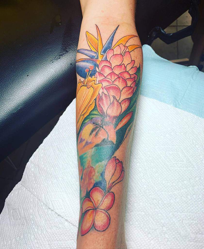 Colorful forearm tattoo featuring a variety of Hawaiian flowers, including a bird of paradise, pink ginger flower, and plumeria, with vibrant leaves and tropical imagery