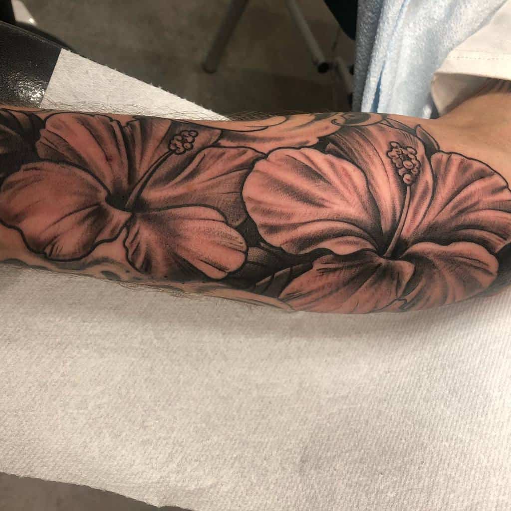 Black and gray forearm tattoo featuring two large, detailed hibiscus flowers with intricate shading and leaf accents