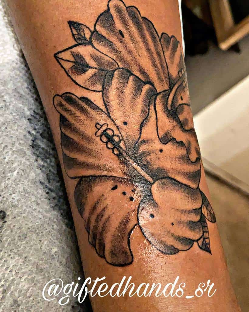 Black and gray forearm tattoo featuring a hibiscus flower with bold outlines, shading, and leaf details