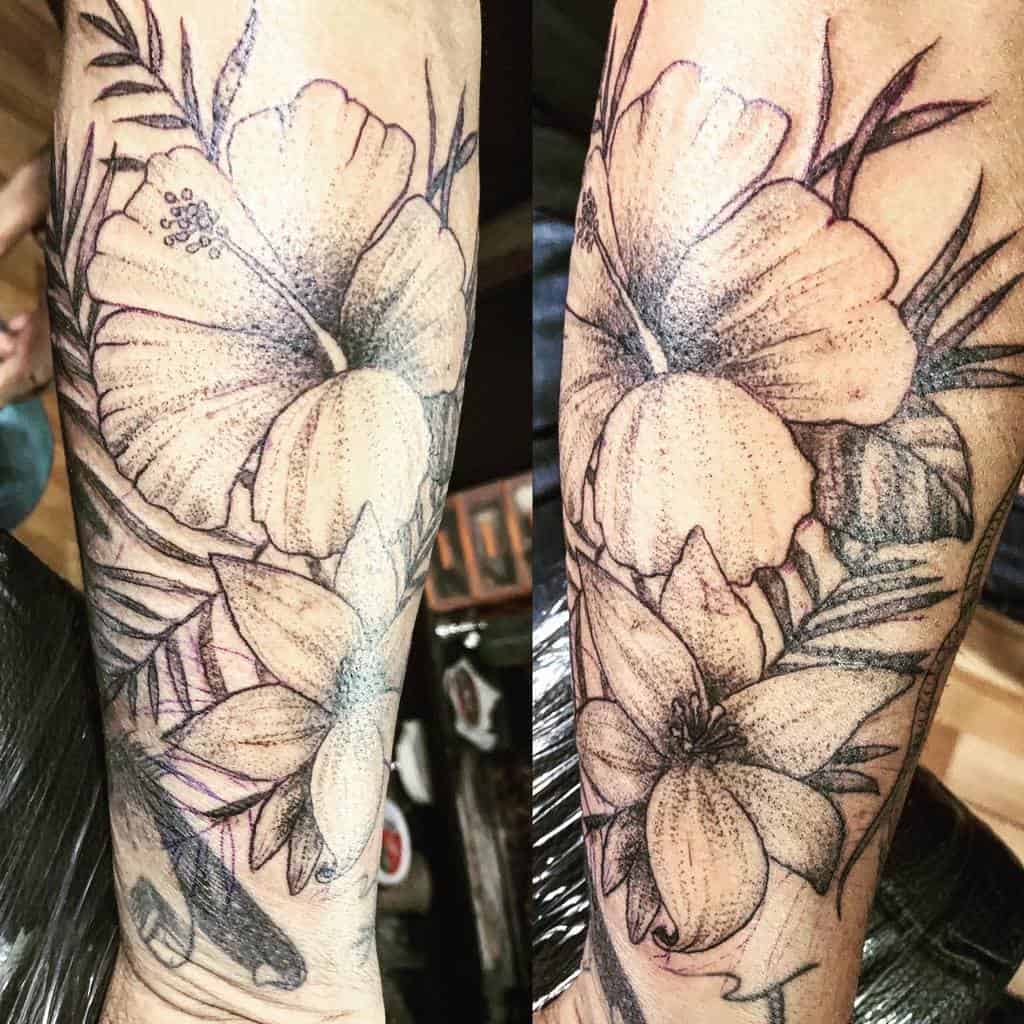 Detailed black and gray forearm tattoo featuring hibiscus flowers and leaves with stippling and shading effects