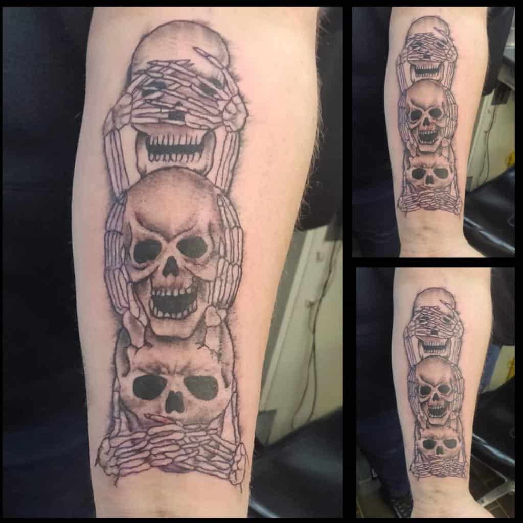 Forearm tattoo of three skulls stacked vertically, with skeletal hands covering eyes, ears, and mouth, symbolizing 'Hear No Evil, See No Evil, Speak No Evil'